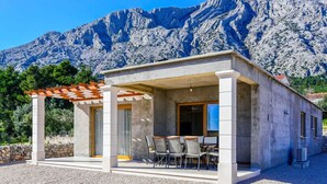 Un área de estar moderna al aire libre con un pérgola, una mesa y sillas, con montañas al fondo.