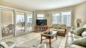 A living room with a sofa, a glass-top coffee table, a television on a wooden cabinet, and a view of the outdoors through large sliding glass doors.