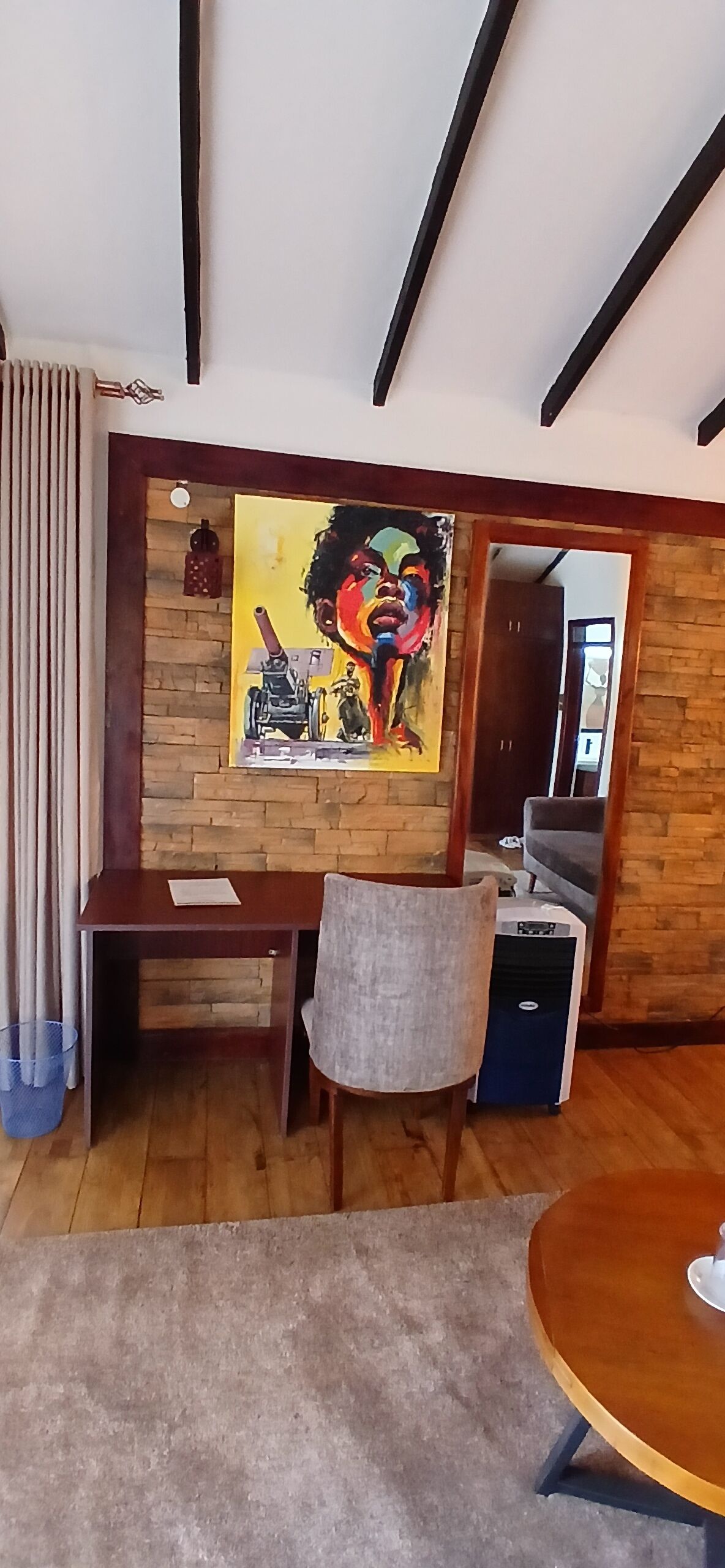 A dining area with a wooden table and chairs, a painting on the wall, and a blue trash bin.