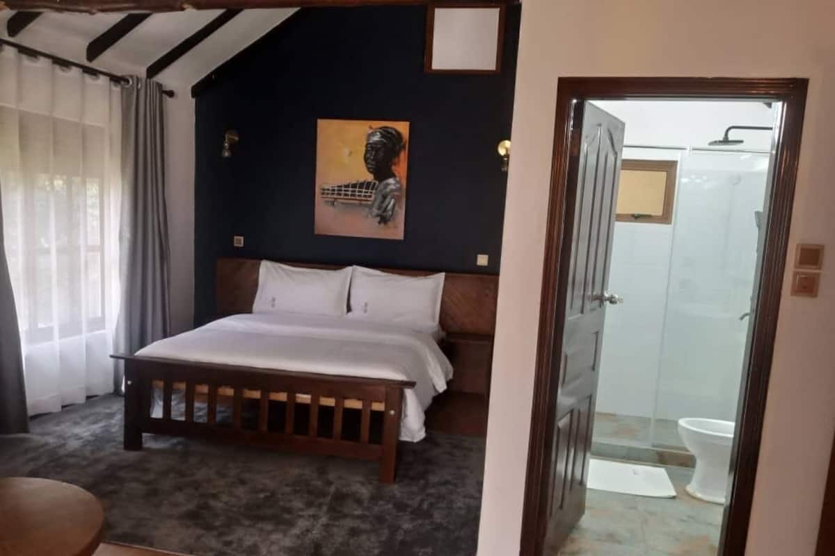 A bedroom with a bed, a wooden headboard, a painting on the wall, a bathroom with a glass shower door, and a small table.