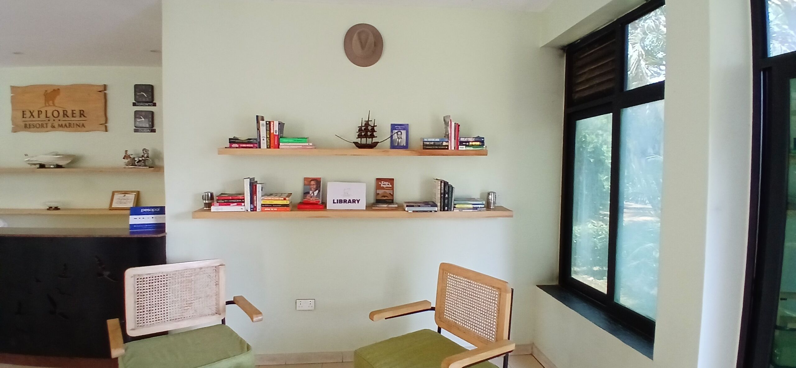 A room with two chairs, a bookshelf with books and decorative items, a sign reading "Explorer Resort & Marina", and a window with a view of trees.