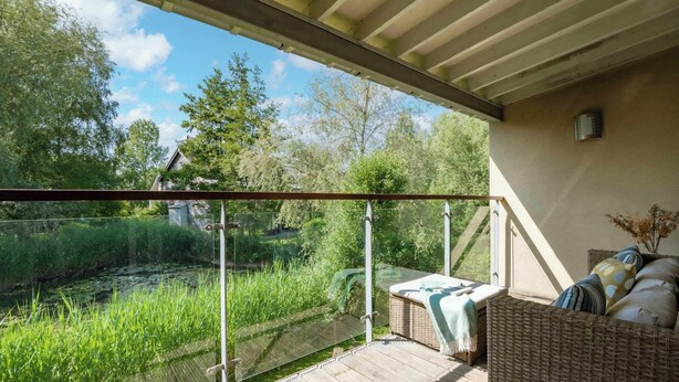 A covered balcony with a glass railing overlooking a garden and a pond. - Beaverbrook Lodge | Family | Spa | Pool Access (Somerford Keynes)