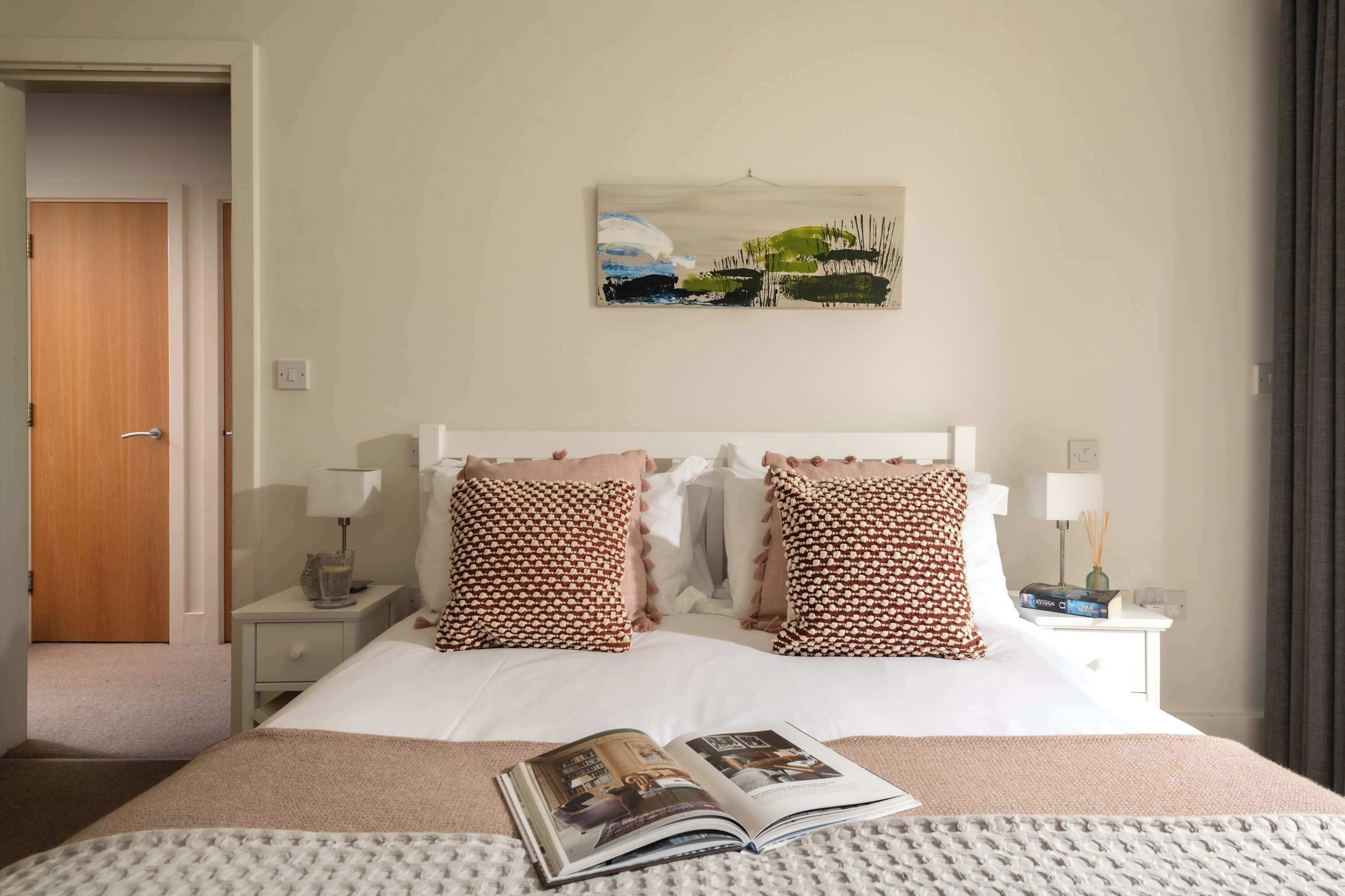 Une chambre d’hôtel bien rangée, avec un lit, une table de chevet, une lampe, un tableau au mur et un magazine sur le lit.