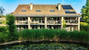 A two-story building with a flat roof, large windows, and a balcony overlooking a body of water with lily pads. - Beaverbrook Lodge | Family | Spa | Pool Access (Somerford Keynes)