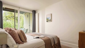 A bedroom with a bed, a bedside table, a glass of water, a book, and a window with curtains. - Beaverbrook Lodge | Family | Spa | Pool Access (Somerford Keynes)