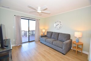 A living room with a grey sofa, two side tables with lamps, a ceiling fan, a wall clock, and a sliding glass door leading to an outdoor deck. - Casa Del Sol 648 3 Bedroom Townhouse (Ocean City)
