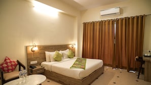 A hotel room with a wooden bed, a nightstand, a desk, a chair, a table, and a window with curtains. - The Dhawala Resort (Alwar)
