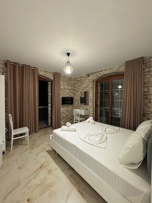 A bedroom with a stone wall, a bed with white linens, a desk, and a chair. - The White Troy (Sarande)