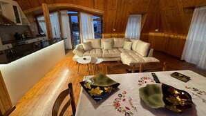 L’intérieur chaleureux d’un chalet avec un plafond en bois, un grand canapé d’angle, une table à manger avec des chaises et un coin cuisine.