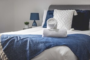 Una cama bien hecha con un edredón azul, almohadas blancas y una toalla enrollada.