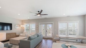 A spacious living room with a grey sofa, a coffee table, a TV mounted on the wall, and a ceiling fan.