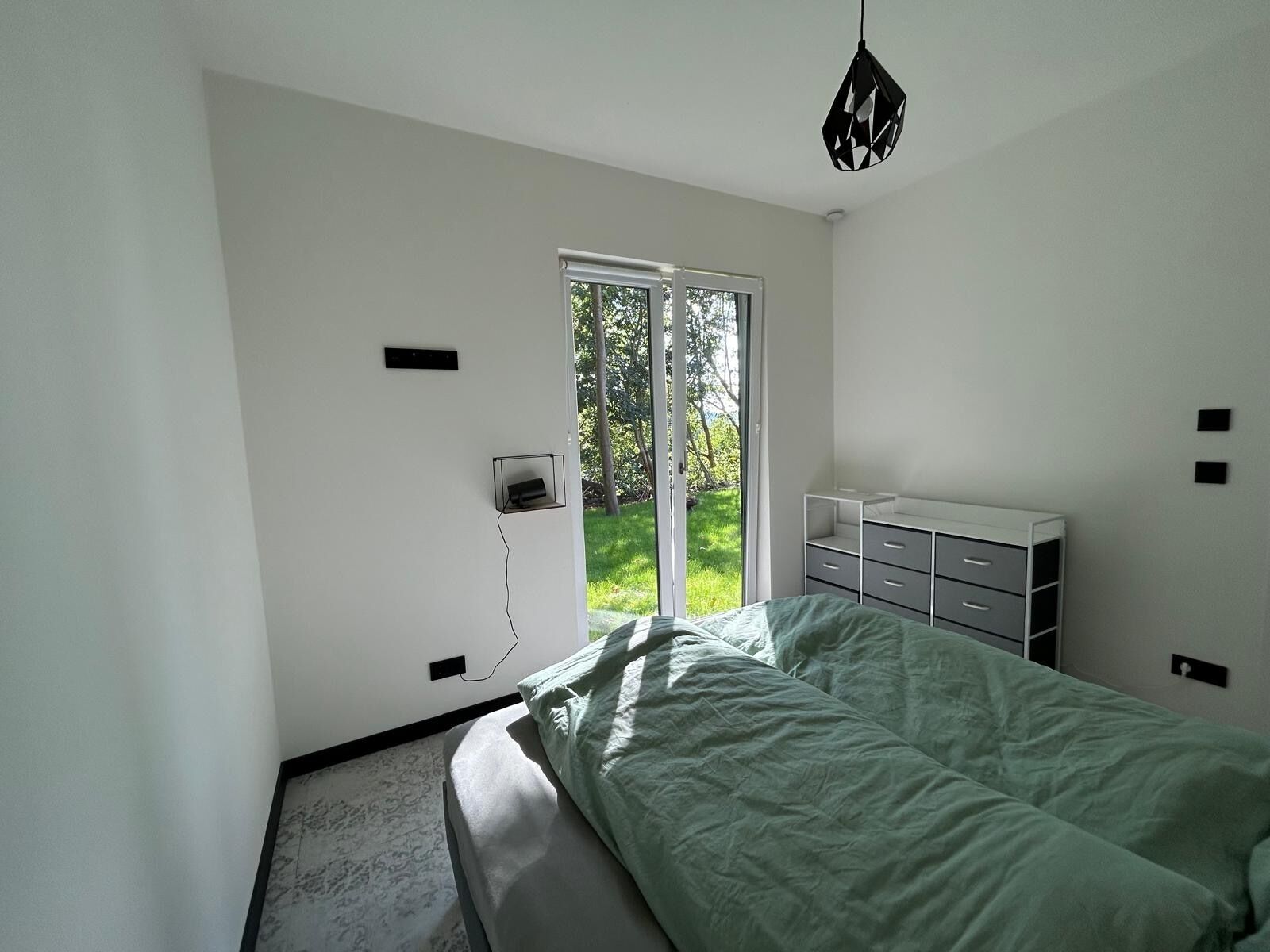 Ein Schlafzimmer mit einem Bett, einer Kommode und einem Schreibtisch. Ein Fenster bietet Blick auf Grünflächen.