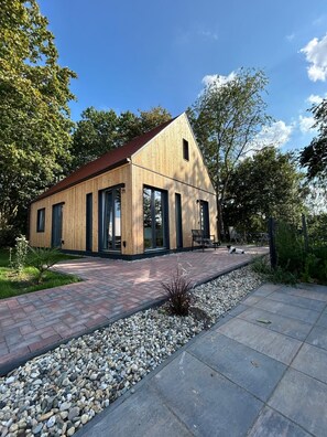 Una casa moderna de madera con ventanas amplias y techo rojo, rodeada de un jardín y un sendero pavimentado.