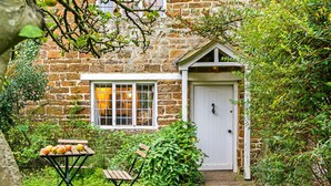 Un bâtiment en pierre avec une porte et des fenêtres blanches, entouré de verdure et agrémenté d’une table en bois sur laquelle sont posées des pommes.