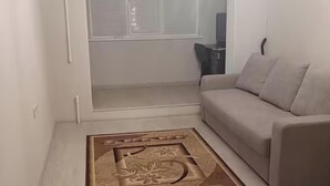 Ein Wohnzimmer mit einem beigefarbenen Sofa, einem gemusterten Teppich und einem Fenster mit Jalousien.