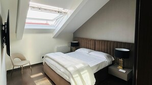 A modern bedroom with a sloped ceiling, a skylight, a bed, a chair, and bedside tables with lamps.