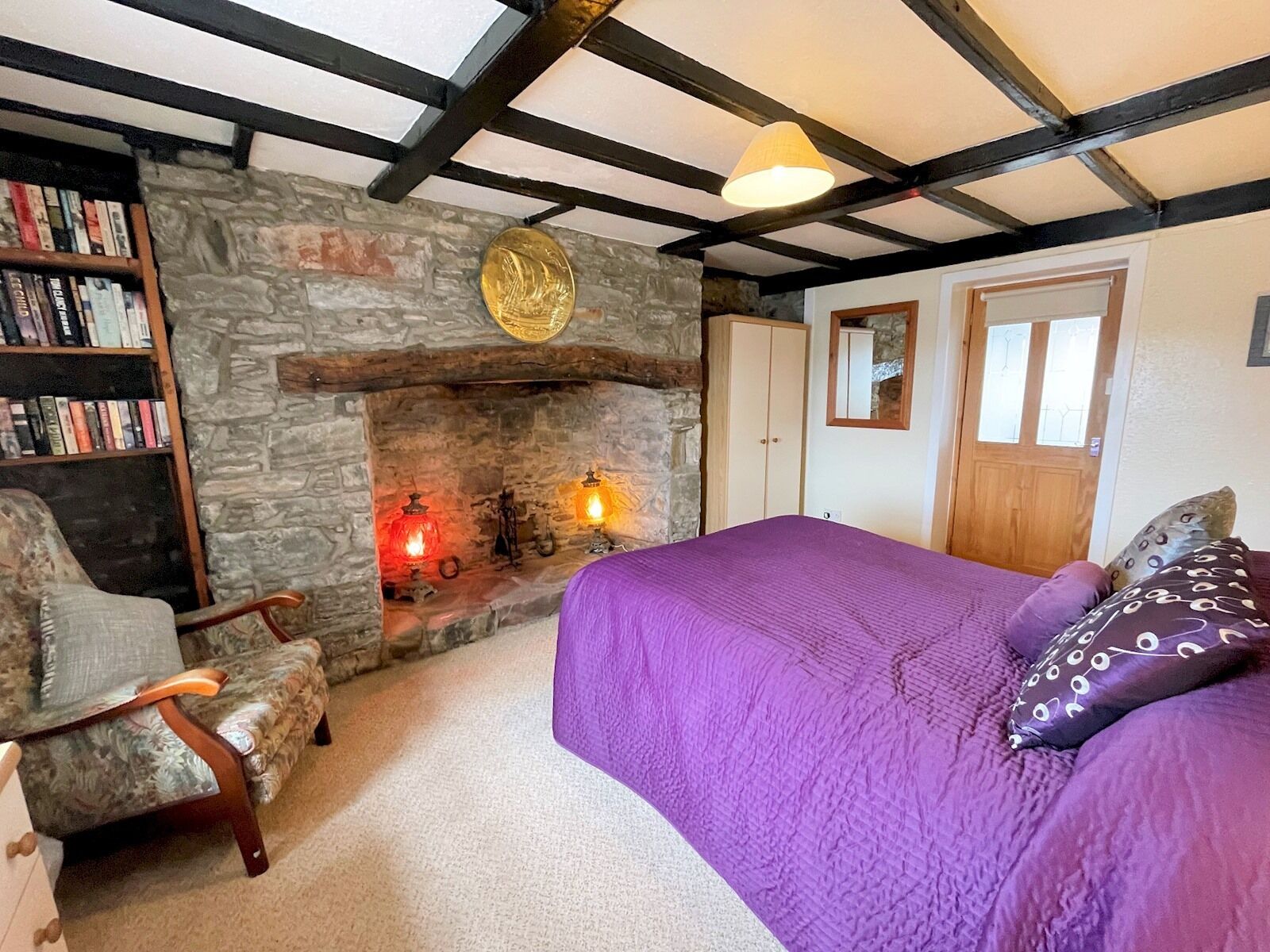A cozy room with a stone fireplace, a bookshelf, a bed with purple bedding, and a wooden chair.
