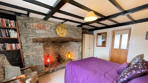 Ein gemütliches Zimmer mit Kamin aus Stein, einem Bücherregal, einem Bett mit violetter Bettwäsche und einem Holzstuhl.