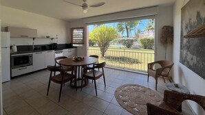 Et moderne køkken med hvide skabe, et spiseområde med et rundt bord og stole, samt en balkon med udsigt til palmer og et hegn.