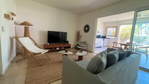 En stue med sofa, fjernsyn, en lænestol, et lille bord og et køkken, der kan ses igennem en åben dør.