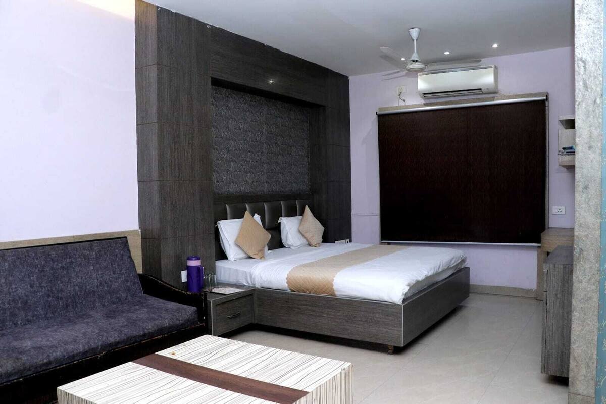 A modern hotel room with a large bed, a sofa, a small table, and an air conditioning unit.