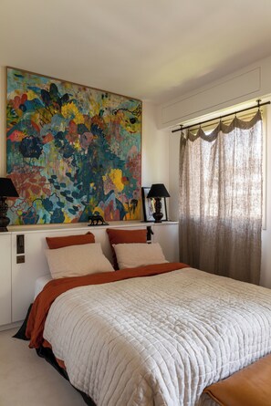 Un dormitorio con un cuadro abstracto grande, una cama con colcha blanca y una ventana con cortinas translúcidas.