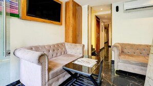 A hotel room with a tufted sofa, a glass table, a wooden cabinet, and a television mounted on the wall.