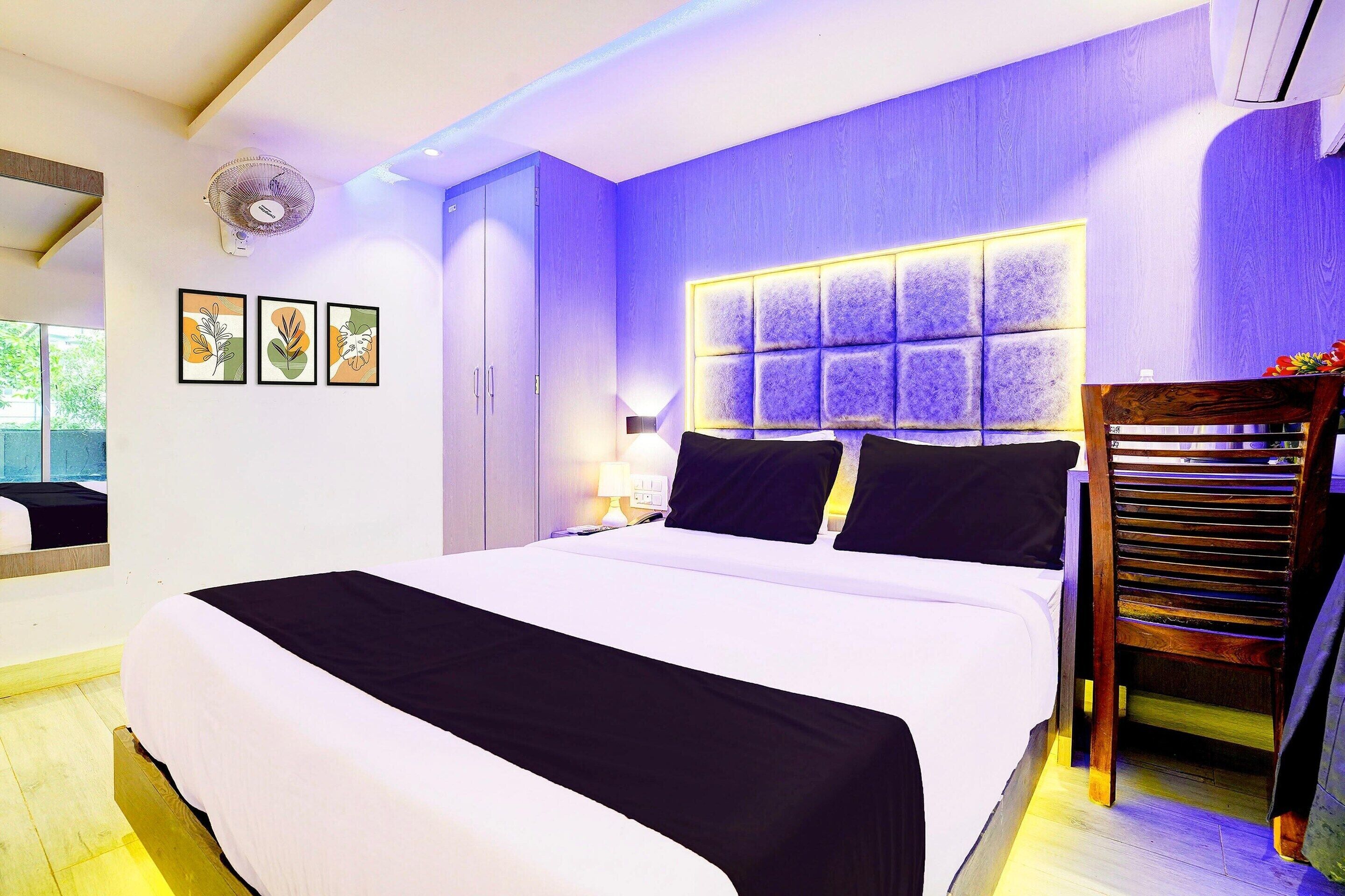 A modern bedroom with a large bed, a headboard with a light panel, a wooden chair, and framed artwork on the wall.