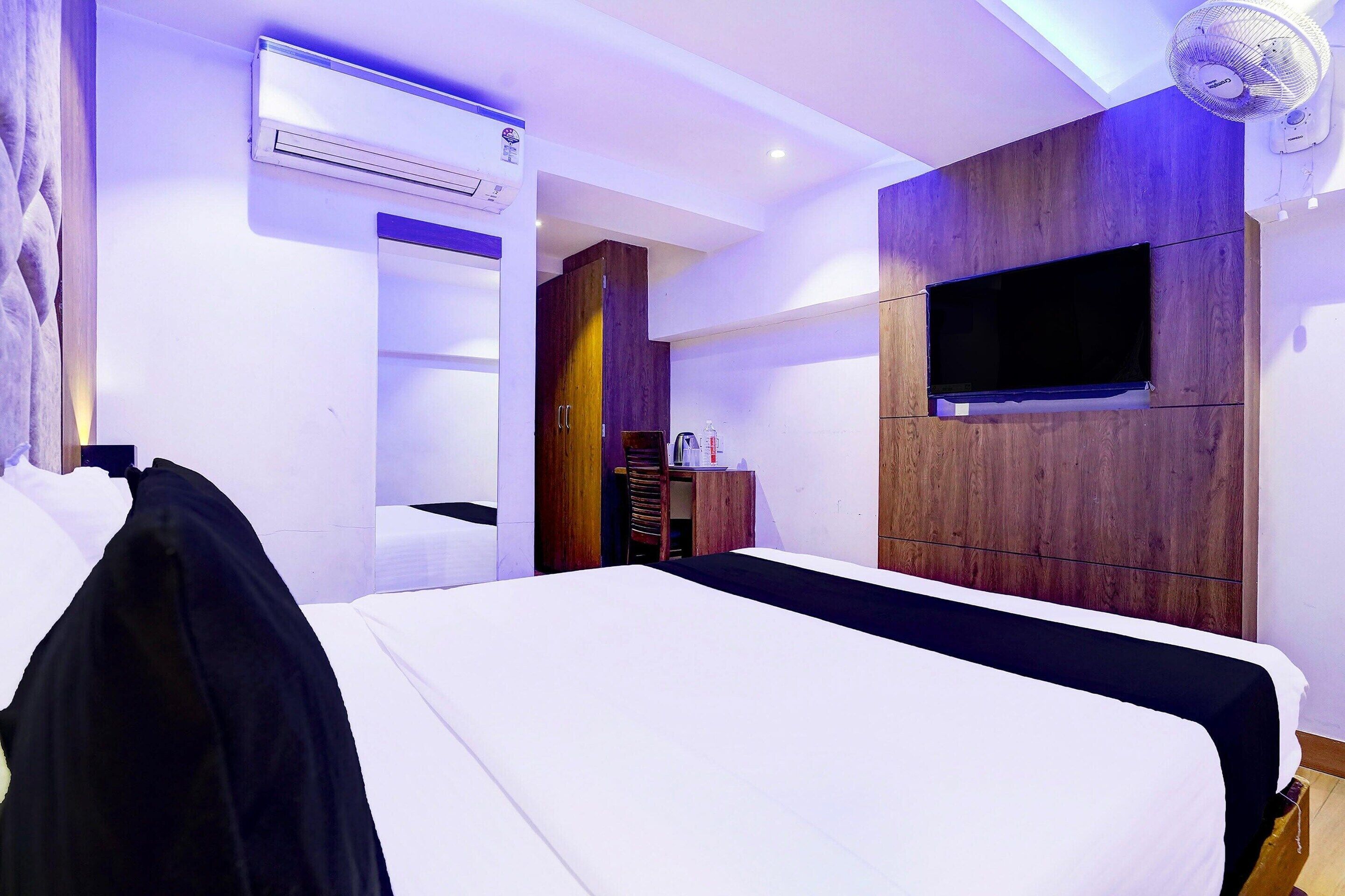 A hotel room with a large bed, a flat-screen TV mounted on the wall, an air conditioning unit, and a wooden wardrobe.