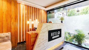 A hotel reception area with a wooden reception desk, a marble counter, a 'HOTEL' sign, and a view of greenery outside.