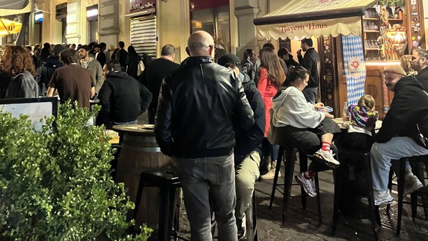 Un café al aire libre concurrido, con mesas y sillas, rodeado de edificios históricos con balcones y señalización.