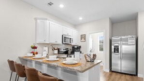 Una cocina moderna con armarios blancos, un refrigerador de acero inoxidable, una encimera de granito y una zona de bar con sillas marrones.