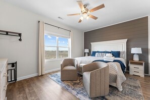 A bedroom with a bed, two chairs, a nightstand, a ceiling fan, and a window with curtains.