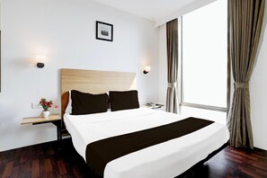 Habitación de hotel con una cama grande, una mesita de noche con lámpara, un jarrón pequeño con flores y una ventana con cortinas.