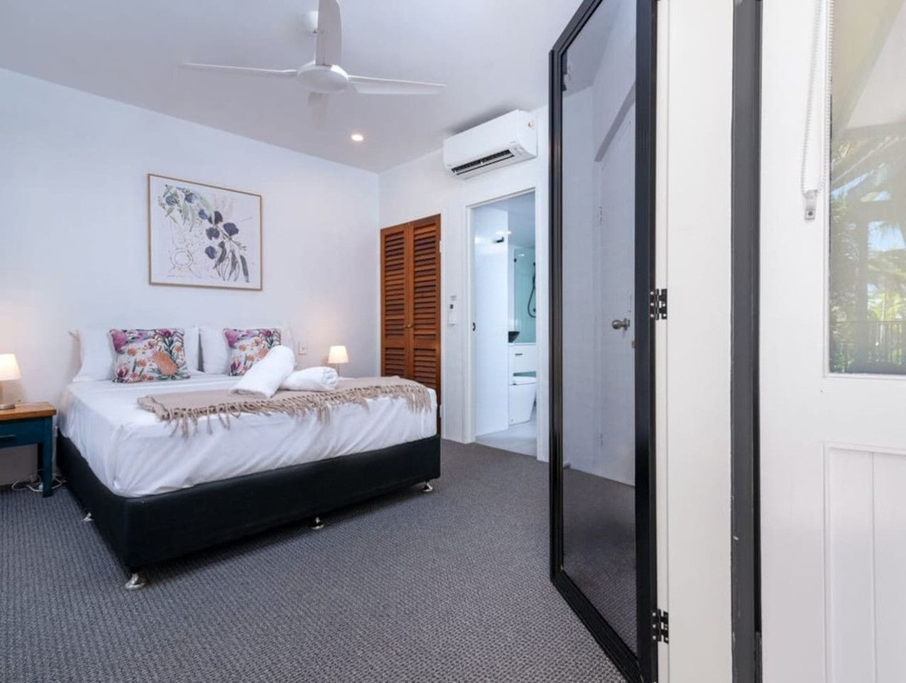 A bedroom with a bed, bedside tables, a ceiling fan, an air conditioning unit, and a door leading to a bathroom.