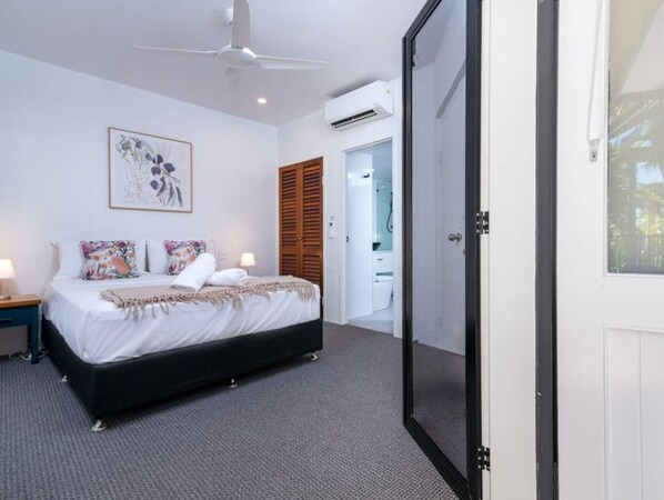 A bedroom with a bed, bedside tables, a ceiling fan, an air conditioning unit, and a door leading to a bathroom. - Cozy-Apartment Near the Beach (Port Douglas)