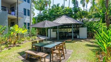 A patio area with a table and chairs, surrounded by lush greenery and a building with a covered seating area.