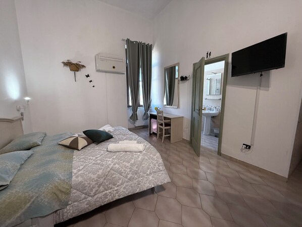 A bedroom with a bed, a desk, a chair, a TV, and a bathroom visible through an open door.
