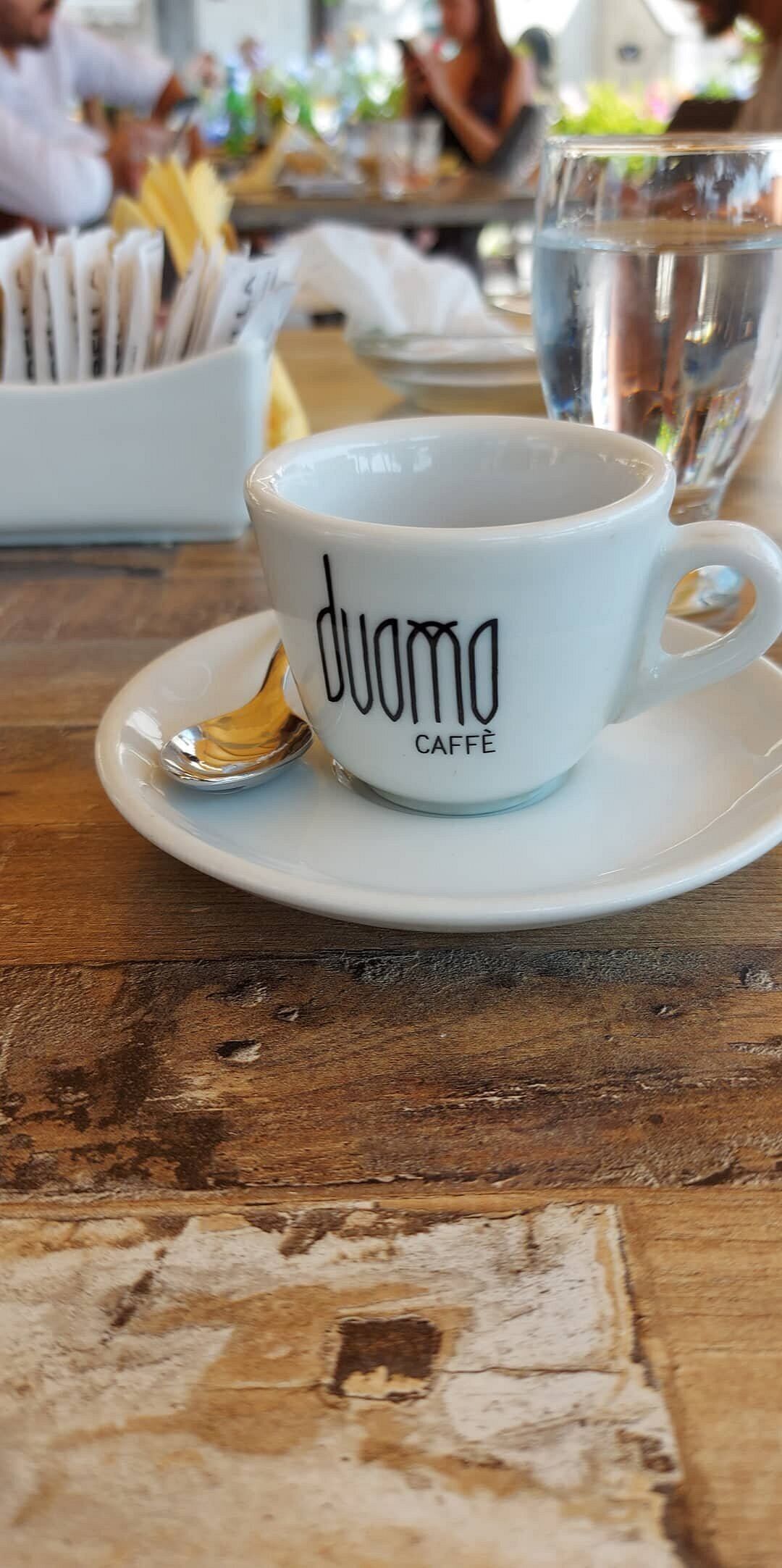 En hvid keramikkop med teksten "duomo Caffè", stående på en underkop med en ske.