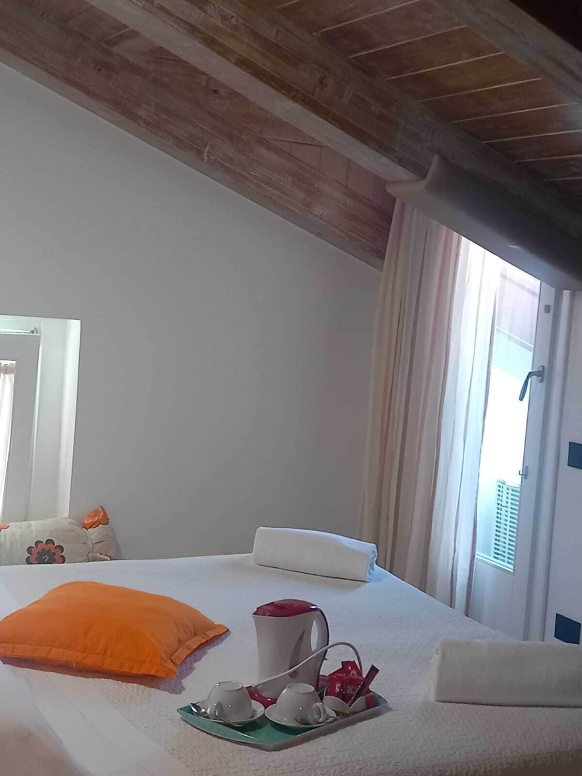 A hotel room with a bed, a window with curtains, a bedside table with a tray of cups and a kettle, and a mirror on the wall.
