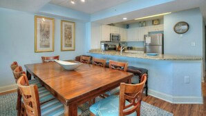 A dining area with a wooden table and chairs, a kitchen with white cabinets and stainless steel appliances, and two framed pictures on the wall. - Grand Atlantic 1407- Oceanfront - Indoor/Outdoor Pools (Myrtle Beach)
