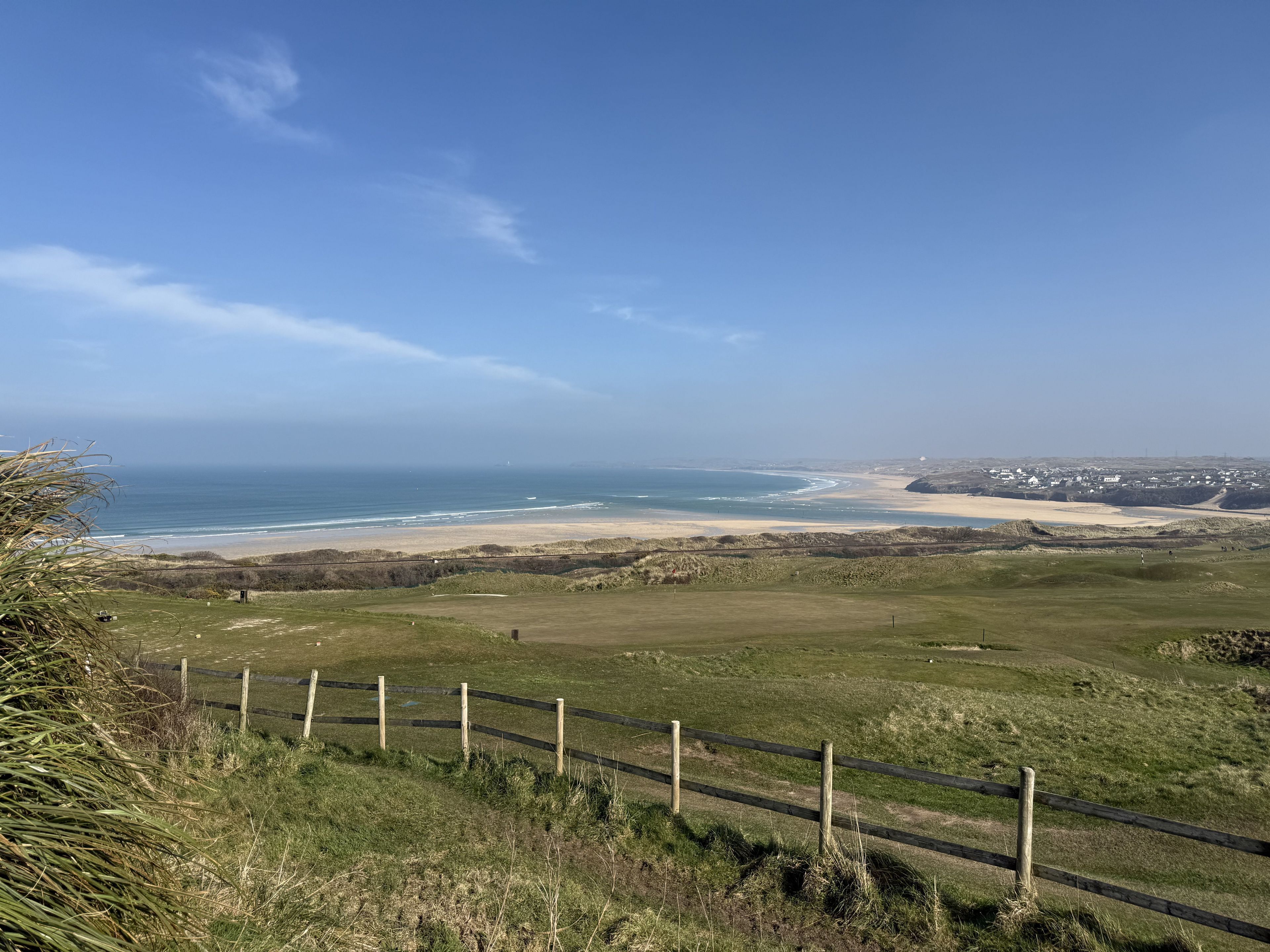 A coastal landscape with a grassy area, a wooden fence, and a distant beach.