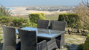 Une table et des chaises en osier, surmontées d’un plateau en verre, offrent une vue sur un paysage côtier avec des maisons et une plage.