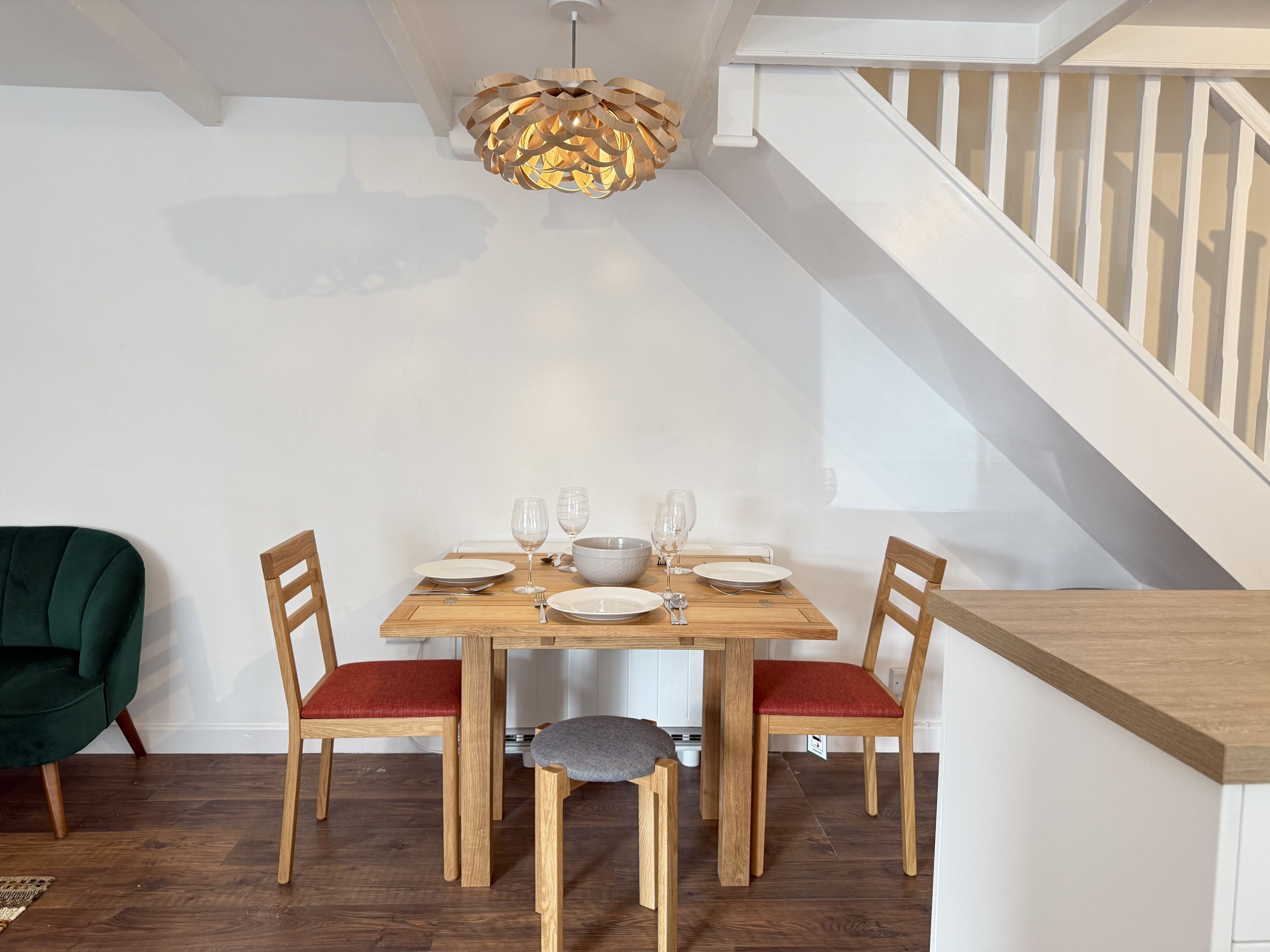 Un comedor con una mesa de madera dispuesta para cuatro personas, dos sillas con cojines rojos, una lámpara colgante de madera original y una escalera con barandillas blancas.