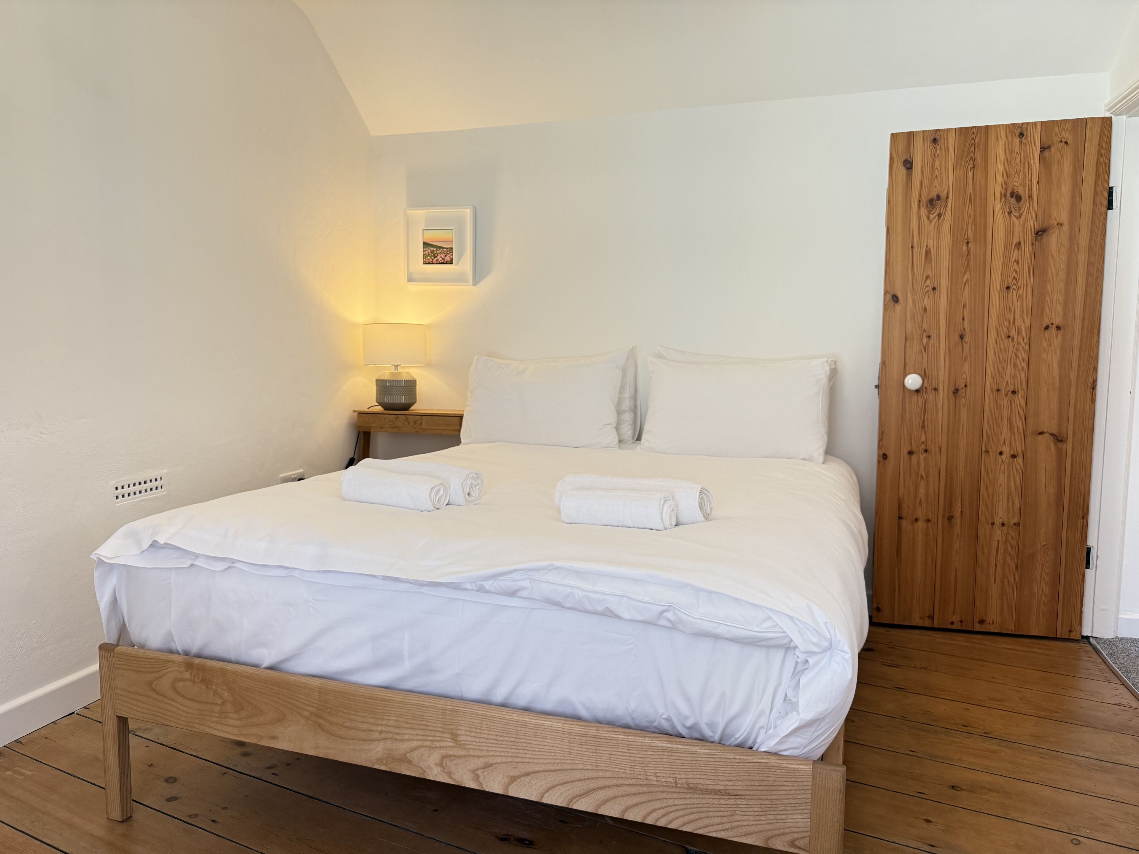 A neatly made bed with white linens and two rolled towels, a bedside lamp, and a wooden door.