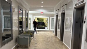 A hotel corridor with a patterned floor, a man walking with a cane, and an exit sign.
