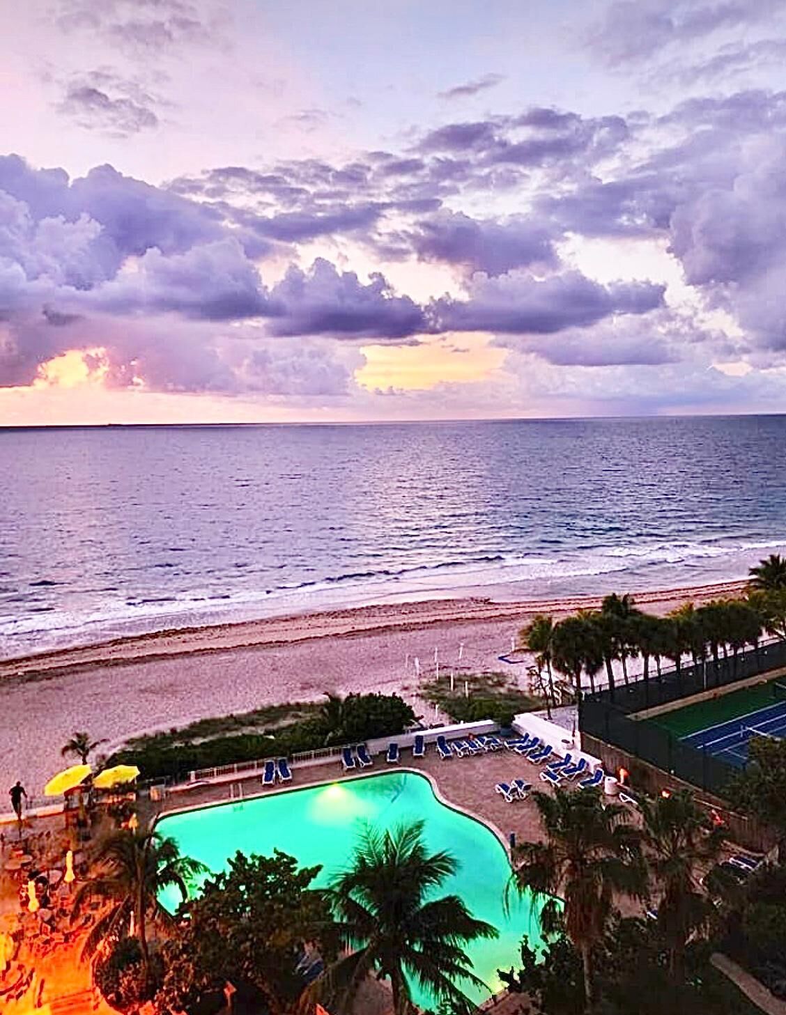 Vista de la playa con piscina, palmeras y cancha de tenis.