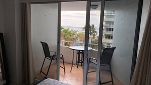 A balcony with a table and two chairs, a bed, and a view of the ocean and buildings.