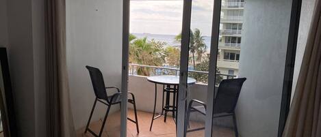 Un balcon avec une table et deux chaises, un lit et une vue sur l’océan et les bâtiments.