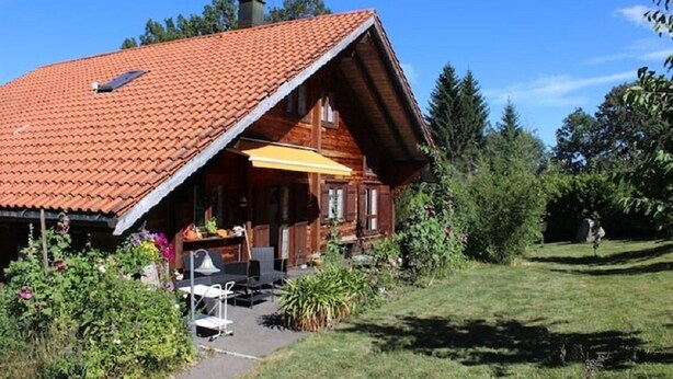 A wooden house with a sloped red-tiled roof, a covered patio with outdoor furniture, and a garden area.